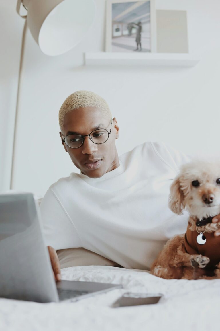 A man in glasses and a white top, with a cute little dog, lays on a bed looking at his laptop.