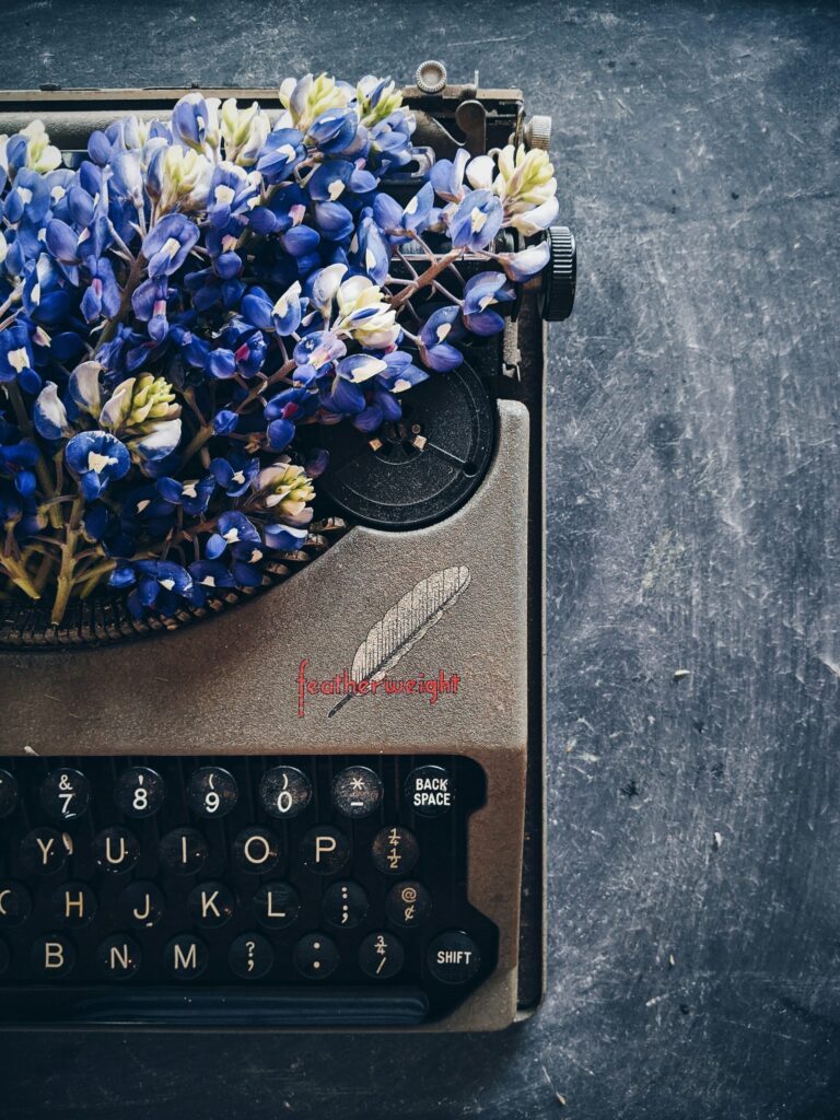 A gray typewriter with purple flowers coming out of its top.