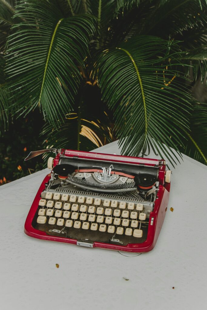 A red typewriter on a white table, with green palm leaves hanging over it.