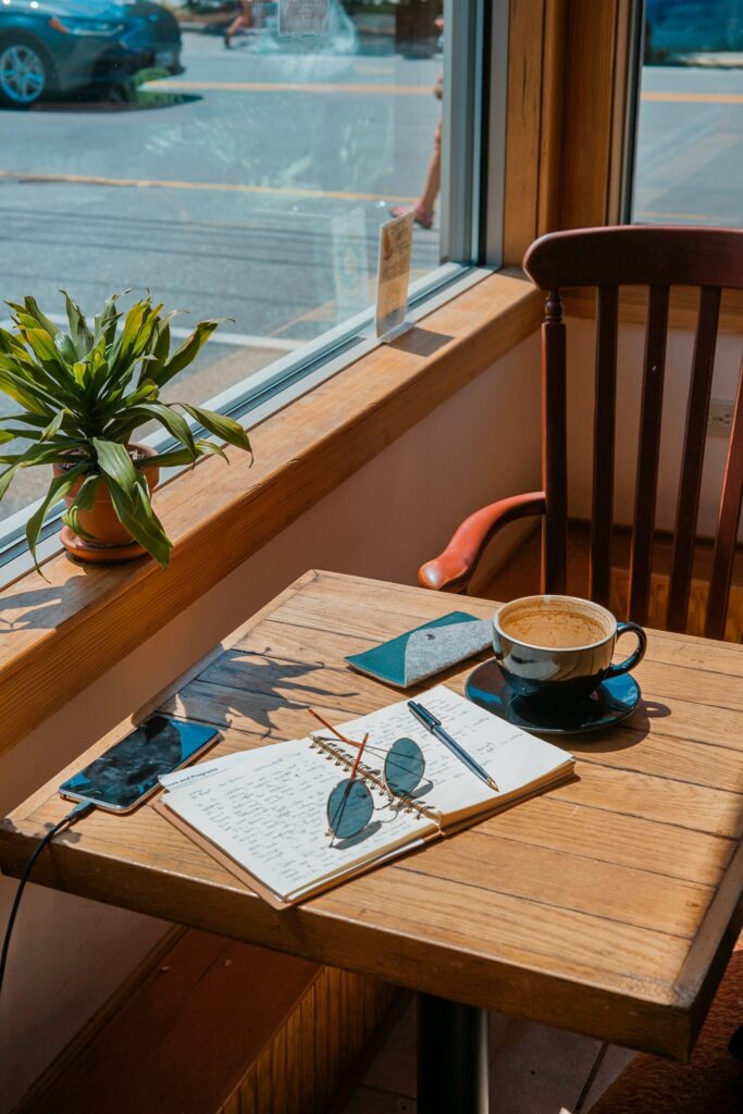 A table by a window contains a journal, a phone, a cup of coffee, and a pair of sunglasses.